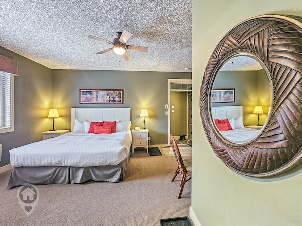 A bedroom with a bed, two bedside tables with lamps, a ceiling fan, a door, and a mirror on the wall. - Golf Course Studio Retreat (Radium Hot Springs)