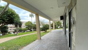 Un paseo techado con baldosas de piedra, la entrada de un edificio y vistas a un área ajardinada con árboles y una carretera.