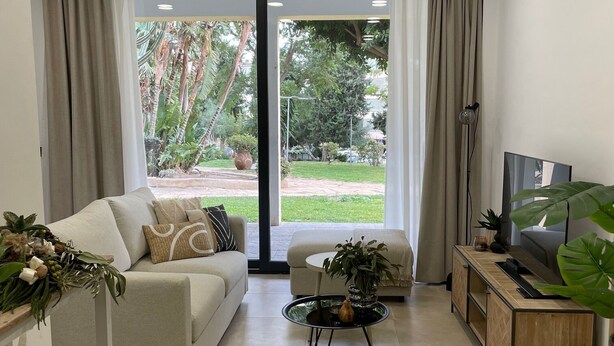 Una sala de estar moderna con sofá, mesa de centro, mueble para TV y ventanas grandes con cortinas.