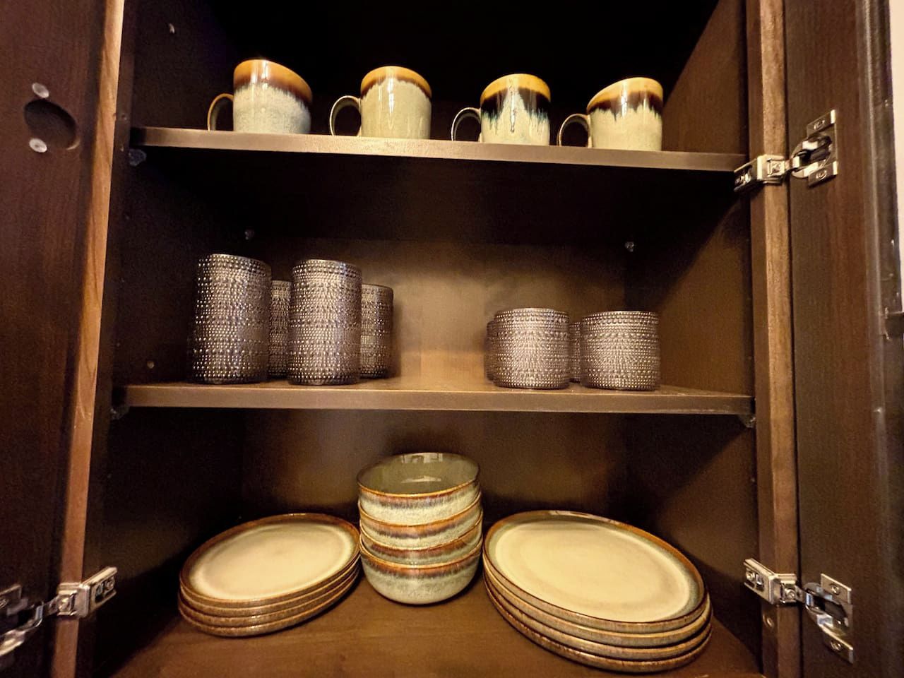 A wooden cabinet with shelves holding ceramic mugs, textured vases, and stacked plates.