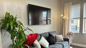 A living room with a grey sofa, a large potted plant, a flat-screen TV, and a floor lamp. - Marilyn Monroe Vintage Suite (Logan)