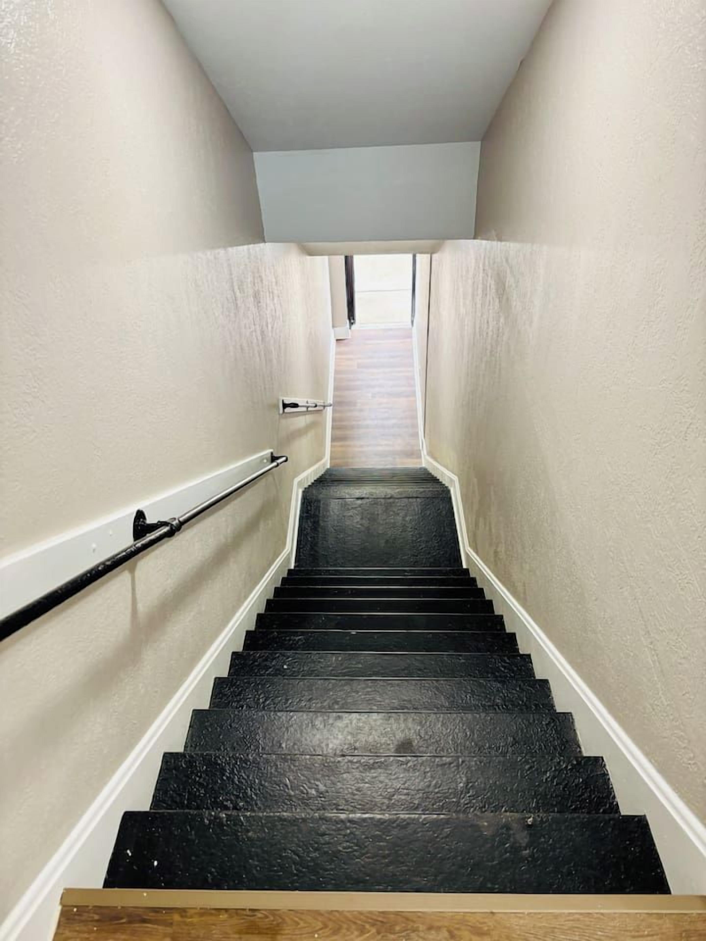 Un escalier étroit avec des marches noires et des murs blancs, menant à un palier où se trouve une porte en haut.