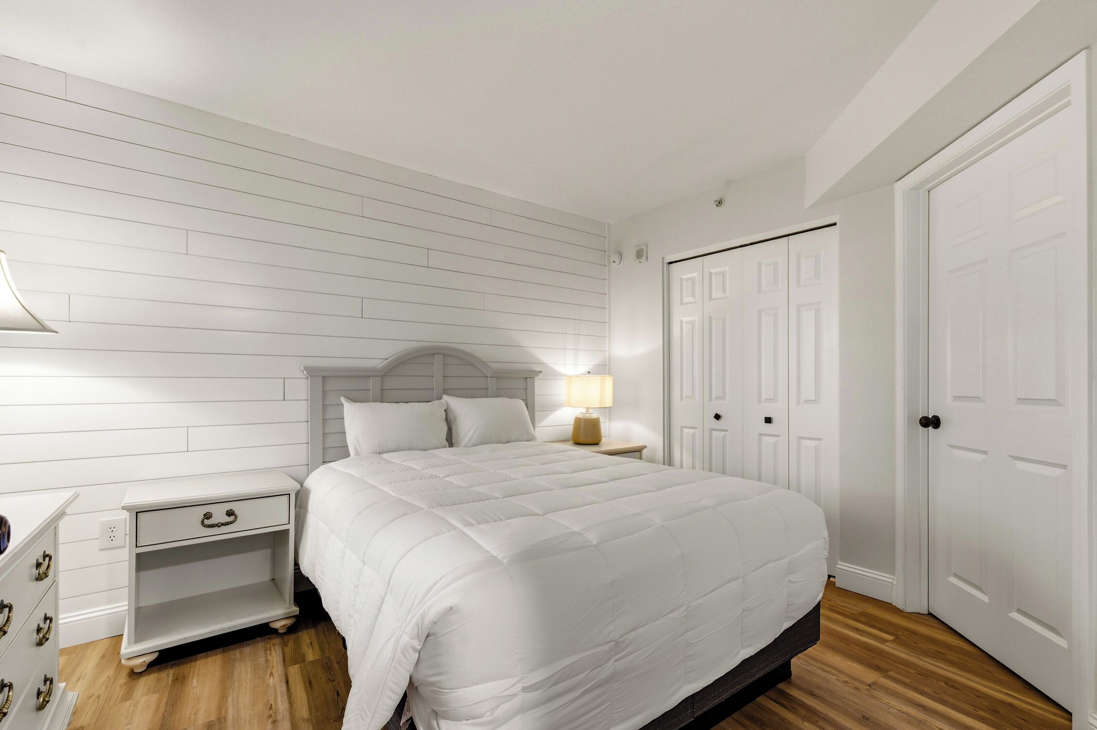 Une chambre à coucher avec un lit blanc, une table de chevet avec une lampe et une armoire.