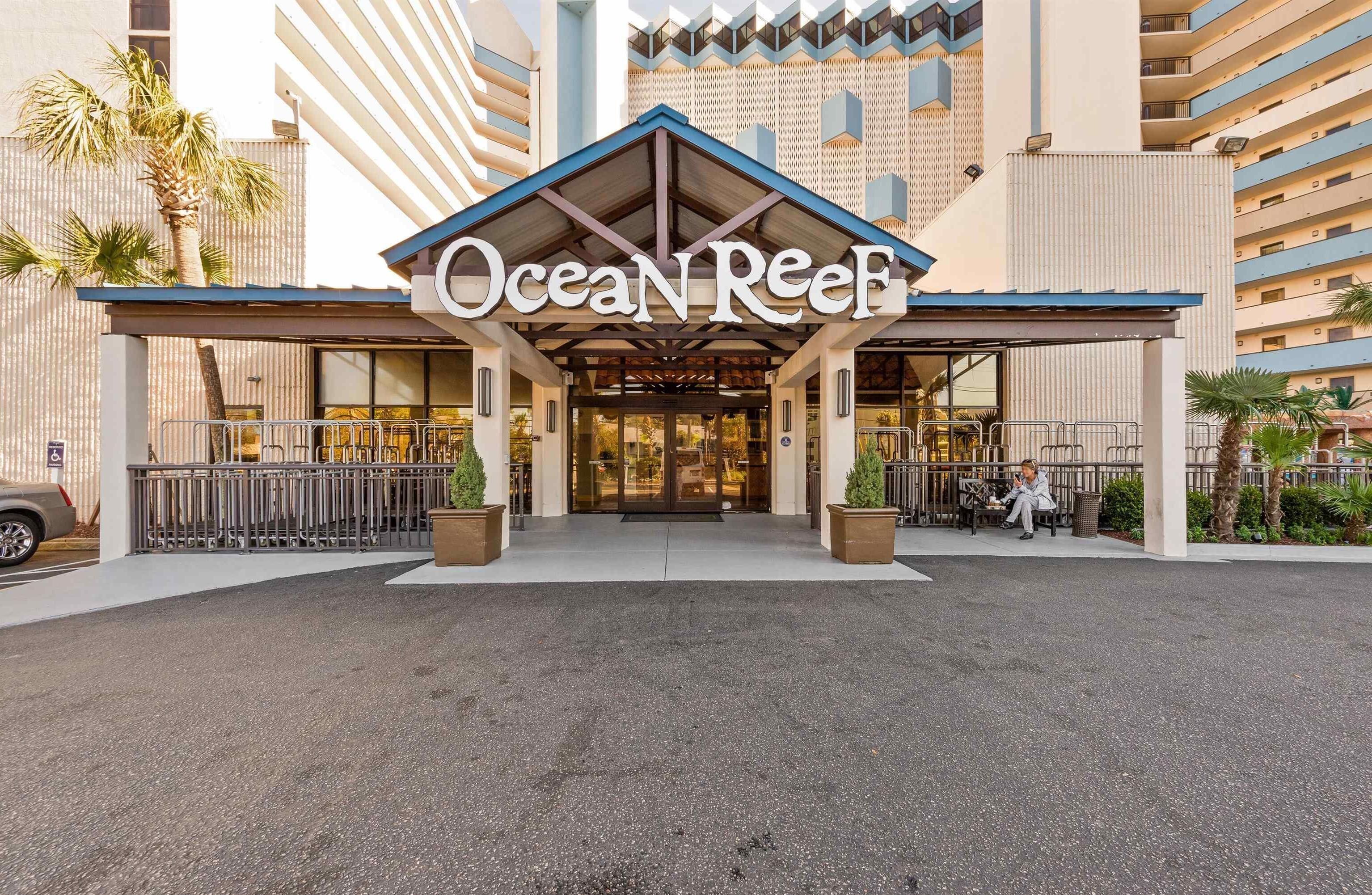 The entrance of Ocean Reef hotel with a covered entrance, a palm tree, and a person sitting on a bench.