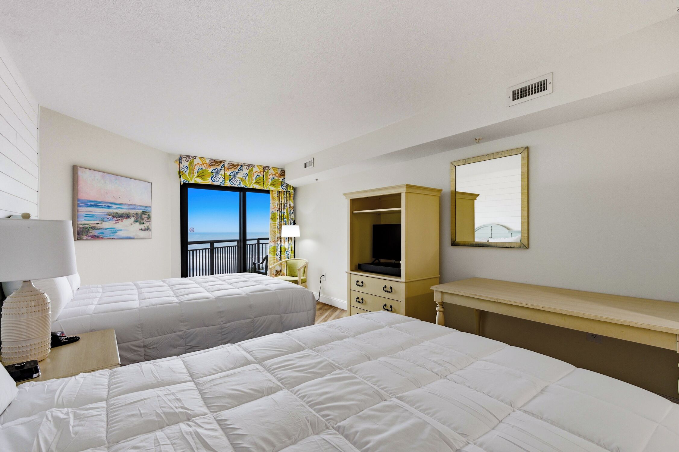 A hotel room with two beds, a TV, a desk, a mirror, and a window with a view of the sea.