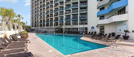 A hotel with a large outdoor swimming pool, multiple balconies, and lounge chairs.