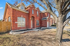 A two-story brick house with a front yard, a wooden fence, and a tree. - Family 4BR Retreat | Pool • BBQ • Game Room (Grand Prairie)