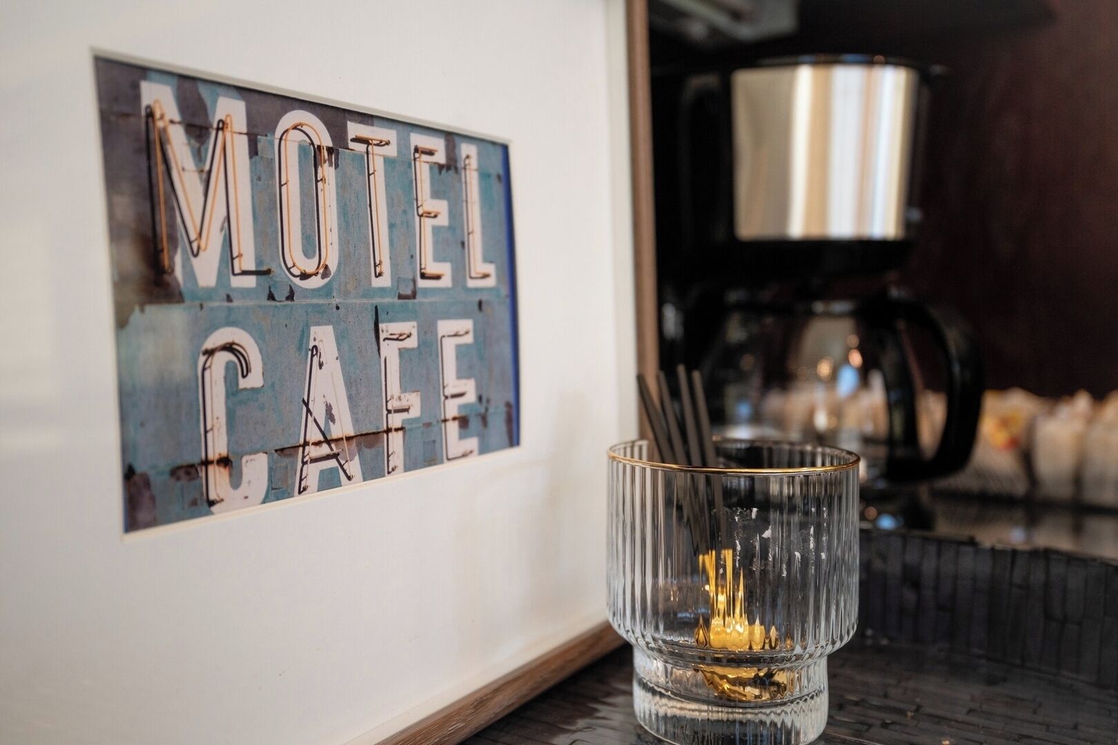 Un letrero enmarcado con la inscripción 'Motel Cafe' sobre una pared, una cafetera de vidrio y un vaso con una bebida sobre una mesa.