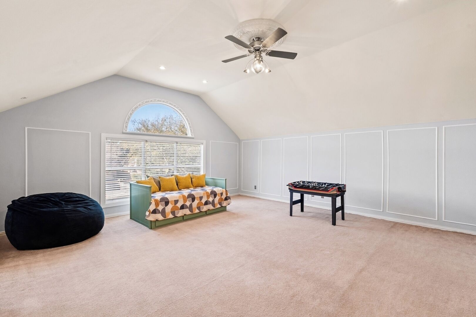 A spacious attic room with a high ceiling, a ceiling fan, a large window with blinds, a bean bag, a sofa with patterned cushions, and a foosball table.