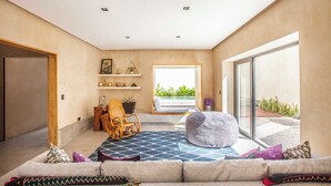 A modern living room with a sofa, a rocking chair, a bean bag, a patterned rug, and a large window with a view of greenery.