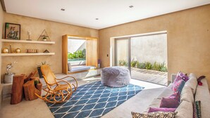 A modern living room with a large window, a wooden rocking chair, a blue patterned rug, and a sofa with cushions.
