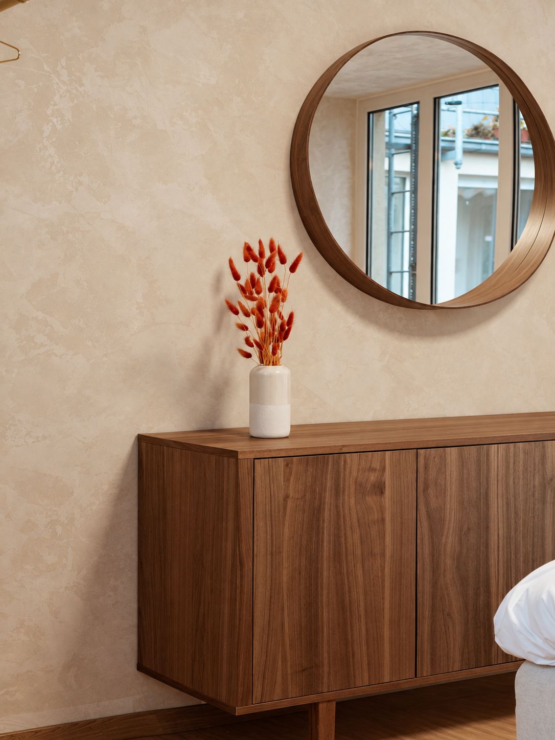 A wooden cabinet with a vase of dried plants on top, a round mirror on the wall, and a window reflecting an outdoor scene.