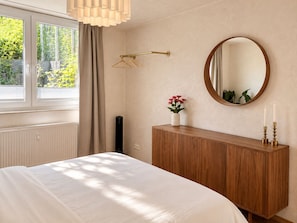 A bedroom with a bed, a wooden dresser, a mirror, a hanging light fixture, and a window with curtains. - Atrium Aachen 2 Apartments (Aachen)