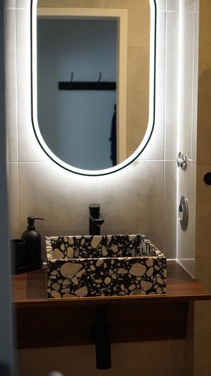 A bathroom with a rectangular sink, a mirror with a light, and a towel rack in the background. - Atrium Apartments Aachen 3 (Aachen)