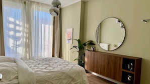 Ein Schlafzimmer mit einem Bett, einer Holzkommode, einem runden Spiegel und einem Fenster mit durchscheinenden Vorhängen.