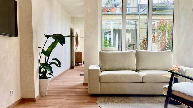 A modern living room with a sofa, a large plant, and a view of a balcony with potted plants. - Atrium Apartments Aachen 1 (Aachen)