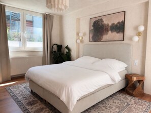 A bedroom with a large bed, a window with curtains, a painting on the wall, and a bedside table. - Atrium Apartments Aachen 1 (Aachen)
