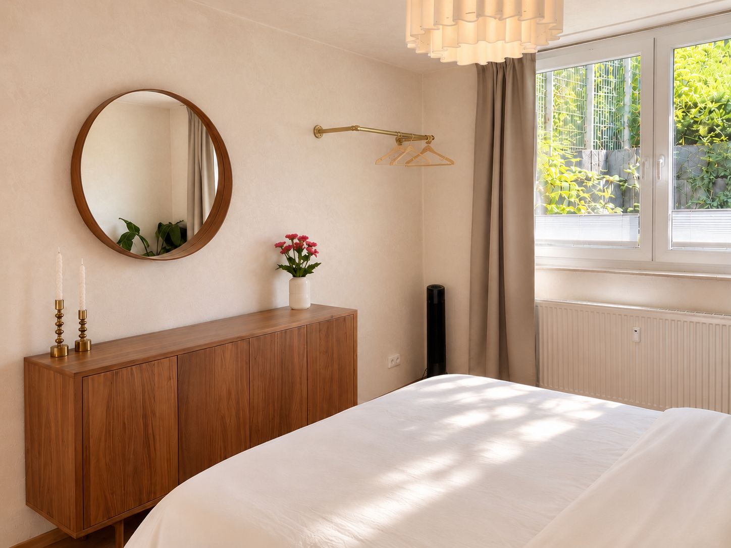 Een slaapkamer met een houten dressoir, een ronde spiegel, een nachtkastje met een bloemenvaas, een raam met gordijnen en een radiator.