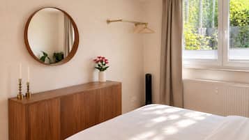 Een slaapkamer met een houten dressoir, een ronde spiegel, een nachtkastje met een bloemenvaas, een raam met gordijnen en een radiator.