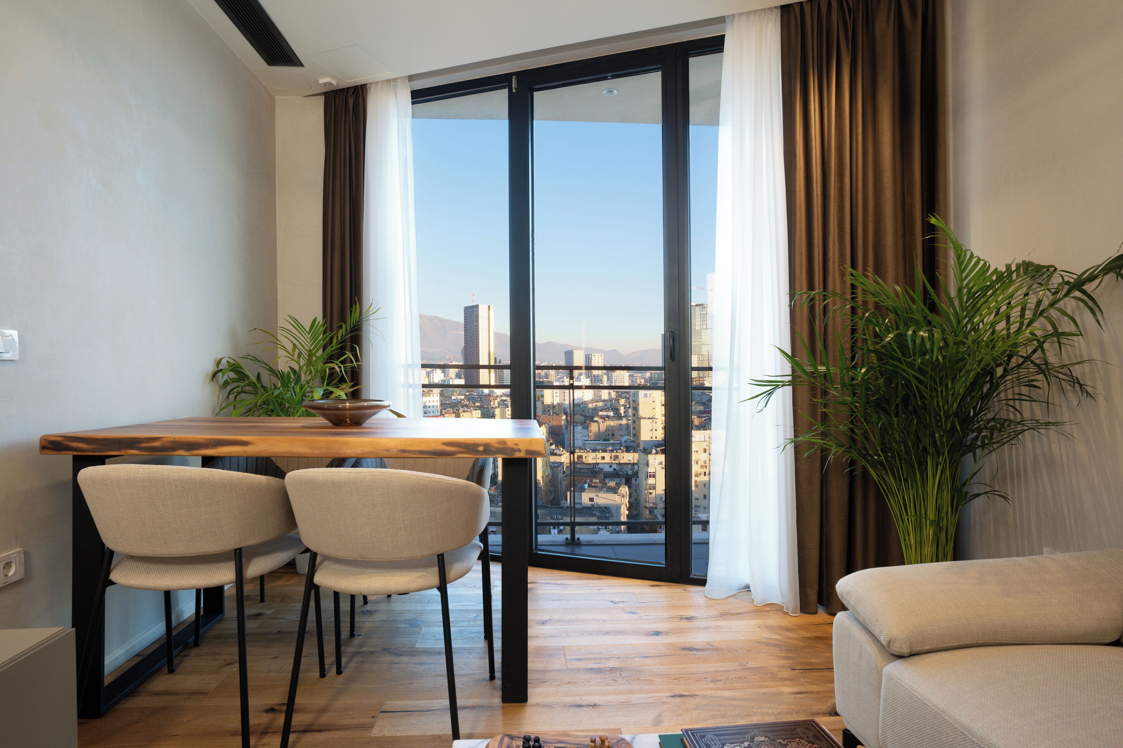 A modern living room with a dining area, large windows with curtains, and a view of a cityscape.