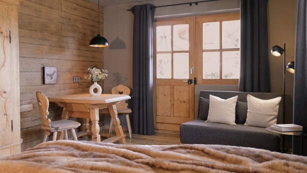 A cozy room with a wooden wall, a dining table with chairs, a sofa with cushions, a window with curtains, and a lamp. - The small apartment (23 square meters) in a modern country house style (Reit im Winkl)