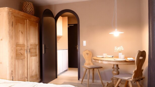 A room with a wooden wardrobe, a round dining table with chairs, and a hanging light fixture. - The newly renovated apartment (30 m²) has a combined living and sleeping area (Reit im Winkl)