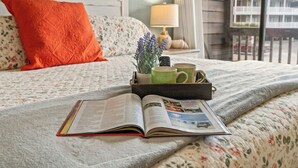 Een keurig opgemaakt bed met een bloemenbedekking, een rood kussen en een dienblad met een boek, twee mokken en een potplant.