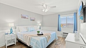 Quarto com cama, mesas de cabeceira, ventilador de teto, janela com cortinas e televisão.