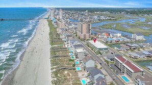 Vista aérea de uma área costeira com praia, edifícios altos e um marcador vermelho indicando a localização.