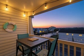 Varanda com mesa e cadeiras, vista para a orla com edifícios ao fundo durante o pôr do sol.