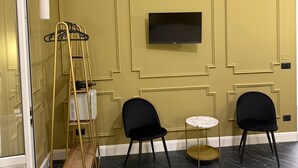 Una habitación con un televisor montado en la pared, dos sillas negras, una mesita redonda y una escalera dorada con repisa.