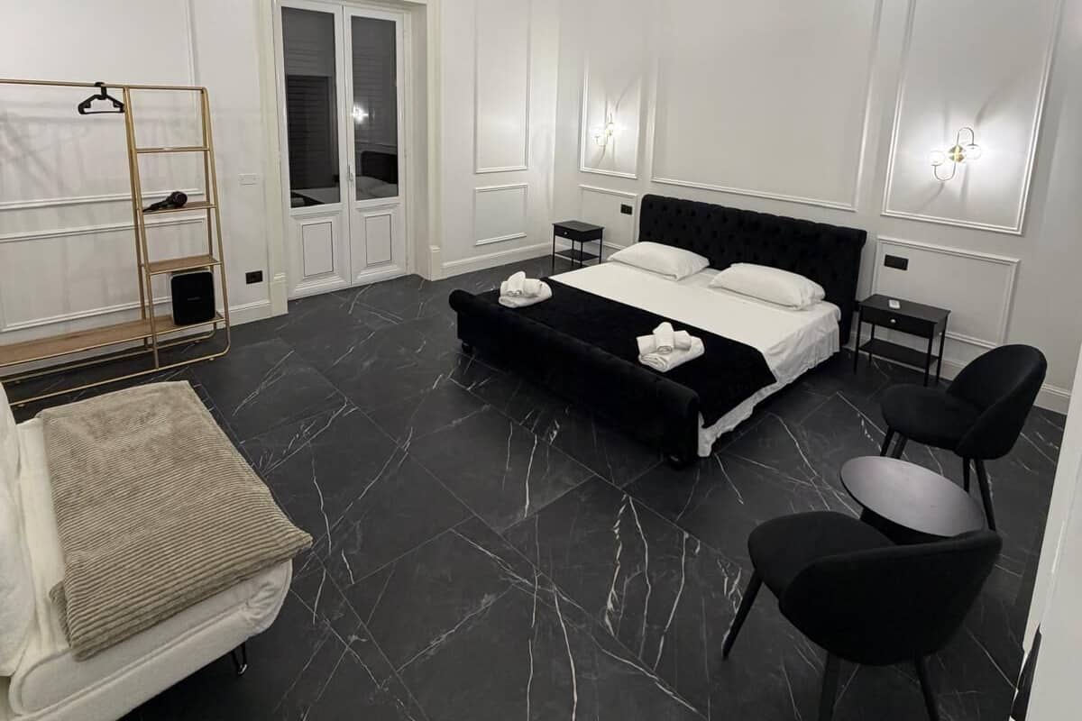A modern bedroom with a black marble floor, a bed with white linens, a black upholstered bench, a gold-framed shelf, and a pendant light.