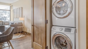 A laundry room with a front-loading washer and dryer, a wooden door, and a seating area with a sofa and chairs. - Charlie's Slopeside: Riverfront Resort/Spa, Ski-In/Out, Private Deck, Mountain Views, Remodeled #821 (Avon)