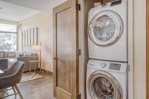A laundry room with a front-loading washer and dryer, a wooden door, and a seating area with a sofa and chairs. - 2BR ski-in/out condo with mountain and pool views in Avon. (Avon)