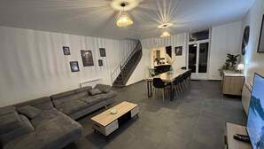 Ein modernes Wohnzimmer mit einem Ecksofa, einem Couchtisch und einem Essbereich mit langem Tisch und Stühlen. Eine Treppe führt zu einer oberen Ebene.