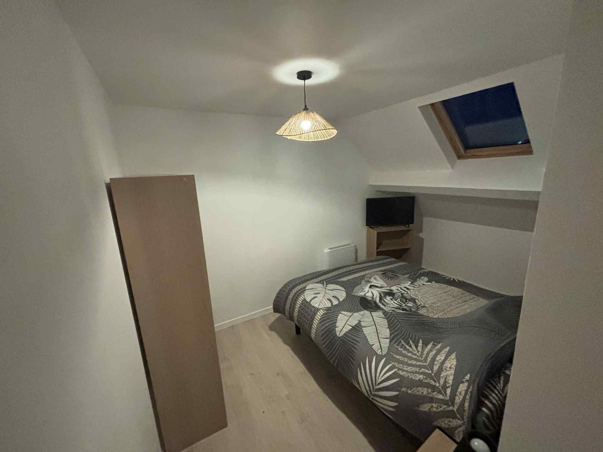 Un dormitorio con cama, luz de techo, televisor sobre un mueble bajo y una ventana pequeña.