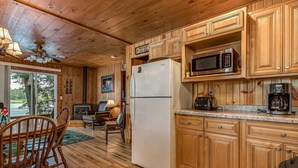 A wooden cabin interior with a kitchen featuring a refrigerator, microwave, toaster, and coffee maker. There is a dining area with a table and chairs, and a large window offering a view of the outdoors. - Timbers - Bouchard Shores Resort (Minocqua)