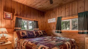 A cozy cabin room with a wooden interior, a large bed with patterned bedding, a nightstand with a lamp, and a window with curtains. - Timbers - Bouchard Shores Resort (Minocqua)