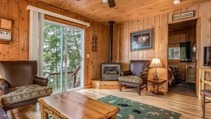 A cozy cabin interior with wooden walls, a fireplace, a large window, and a ceiling fan. - Timbers - Bouchard Shores Resort (Minocqua)