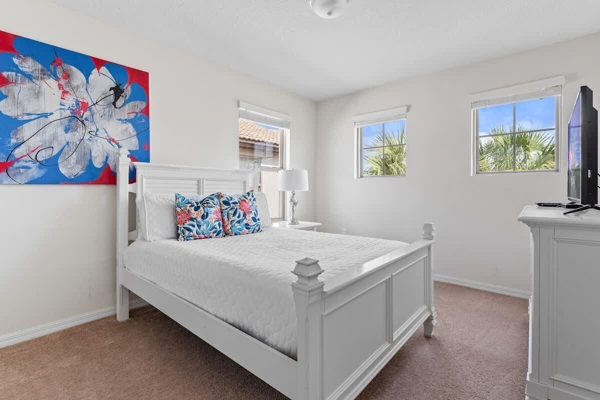 Un dormitorio con cama blanca, mesita de noche, lámpara, televisor sobre un mueble y un cuadro abstracto grande en la pared.