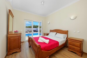 Quarto com cama de madeira, mesa de cabeceira, cômoda e porta de vidro corria para uma piscina.