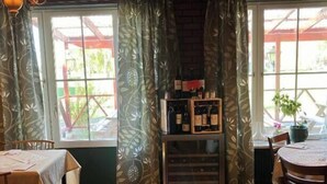 A cozy dining area with wooden tables and chairs, large windows with patterned curtains, and a wine rack against the wall. - Brevensgården (Brevens Bruk)
