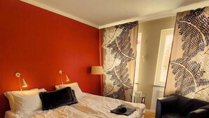 A double bed with white linens, two bedside lamps, and a black chair in a room with red walls and a large fern-patterned curtain. - Brevensgården (Brevens Bruk)