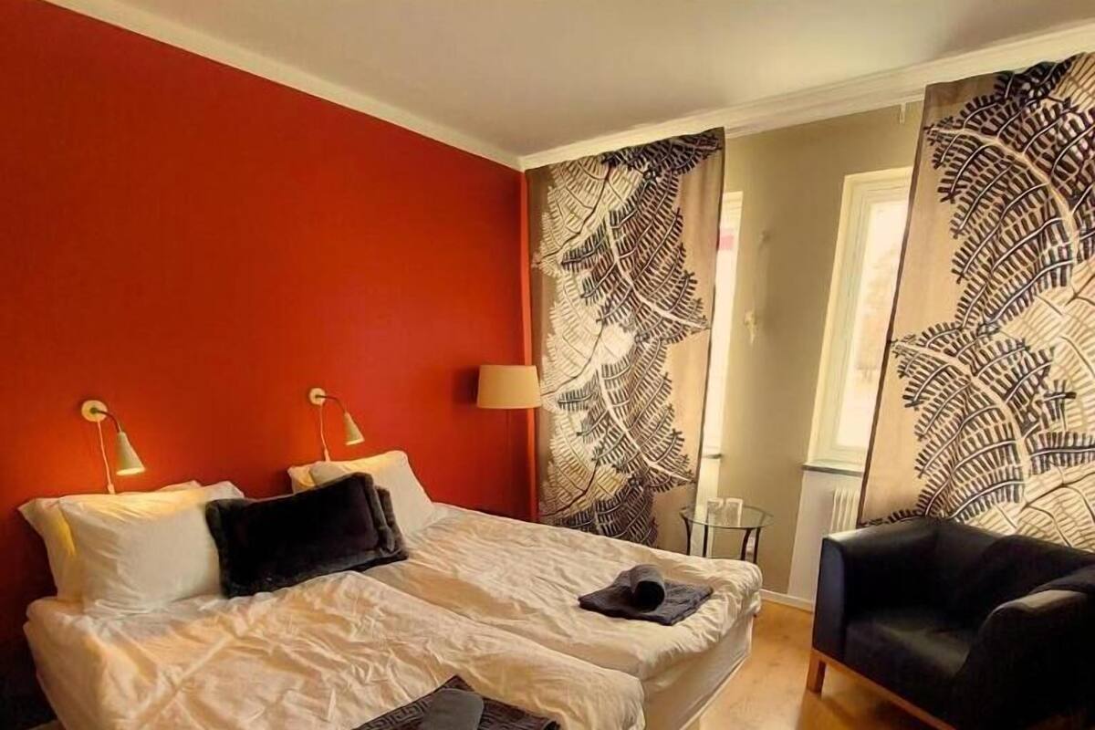A double bed with white linens, two bedside lamps, and a black chair in a room with red walls and a large fern-patterned curtain.