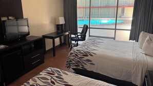 A hotel room with two beds, a desk with a chair, a TV, and a view of a swimming pool.