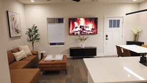 Ein modernes Wohnzimmer mit einem braunen Sofa, einem weißen Couchtisch, einem Fernseher, der einen Kamin zeigt, sowie einem Essbereich mit weißem Tisch und Stühlen.