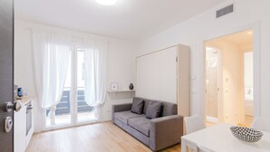 A modern, well-lit living room with a grey sofa, a white table, and a door leading to a balcony. - GINO SUITES - Bilocali Luxury - Appartamenti vicini a Rho Fiera e Milano (Bollate)