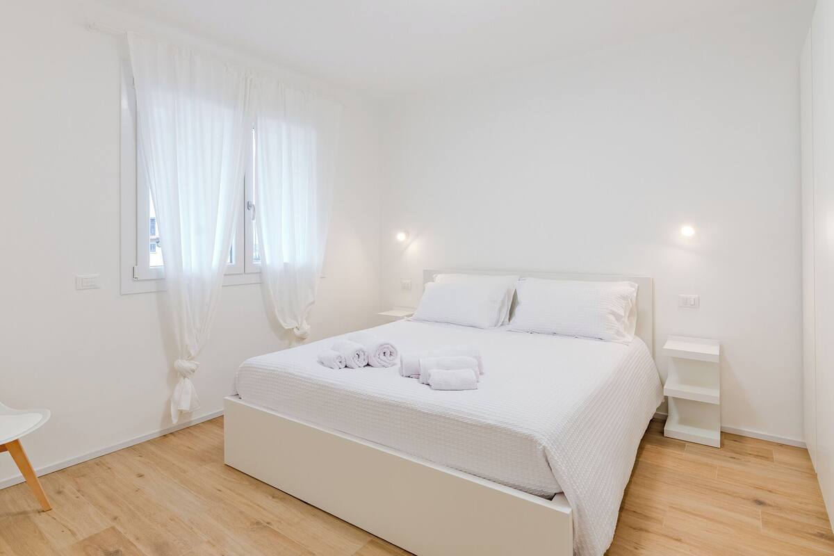 Une chambre à coucher au style minimaliste, avec un lit, des tables de chevet et une fenêtre ornée de rideaux légers.