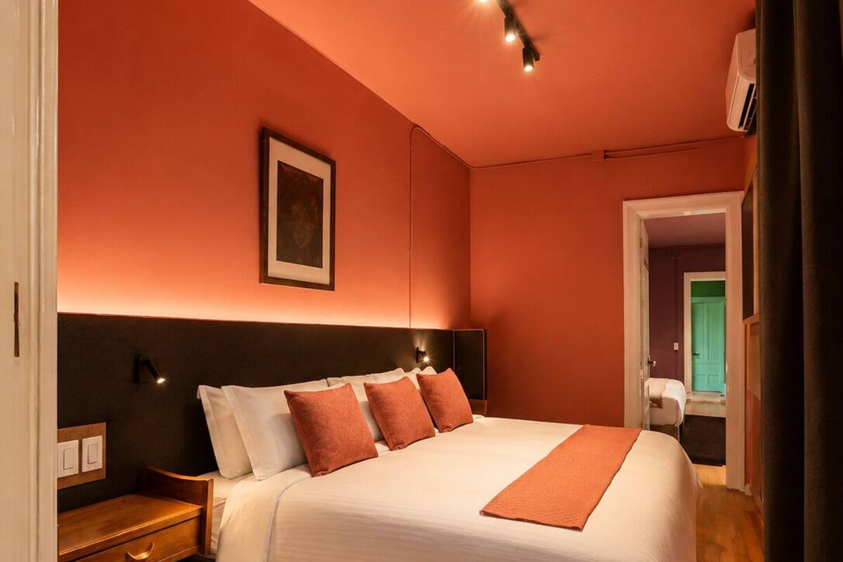Habitación de hotel con una cama grande, una mesita de noche, un cuadro enmarcado en la pared y una puerta que da a otra habitación.