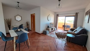 A living room with a sofa, dining table, chairs, and a TV. There's a door leading to a balcony. - Apartment 'Charming Ocean View' with Sea View, Wi-Fi and Air Conditioning (Albufeira)